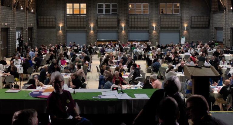 View of the floor of General Synod from the dais. A room full of people around round tables.