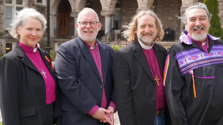 Image of four bishops in purple shirts with clerical collars. They are smiling with a old building blurred in the background.