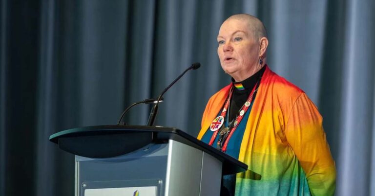 A person with short gray hair wearing colorful rainbow clerical robes speaking at a podium with a microphone.